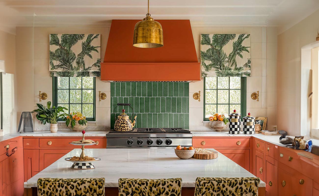 A vibrant kitchen design with a bold orange range hood, green tiled backsplash, and decorative leopard print stools, emphasizing creative interior design solutions.